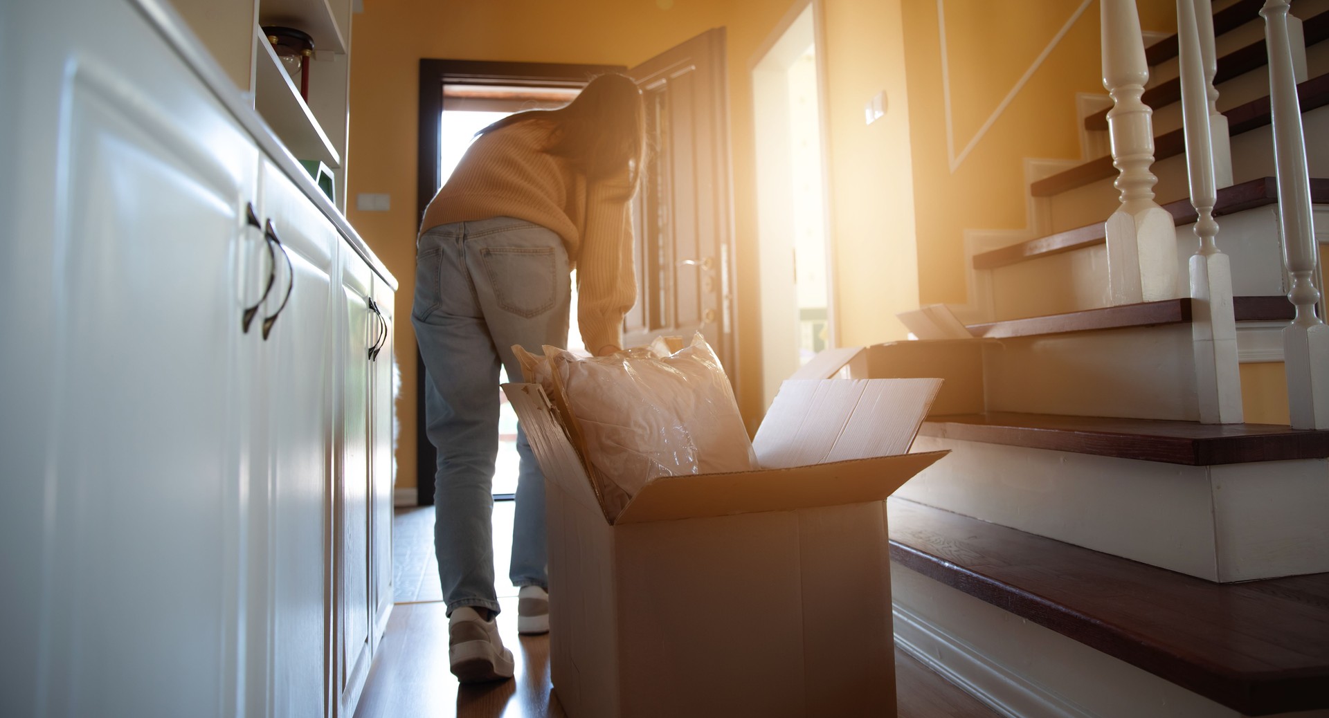 A woman unpacks a moving box in the hallway of her new home, bathed in soft, warm light. The cozy atmosphere and the stairs in the background suggest the start of a fresh and exciting journey.